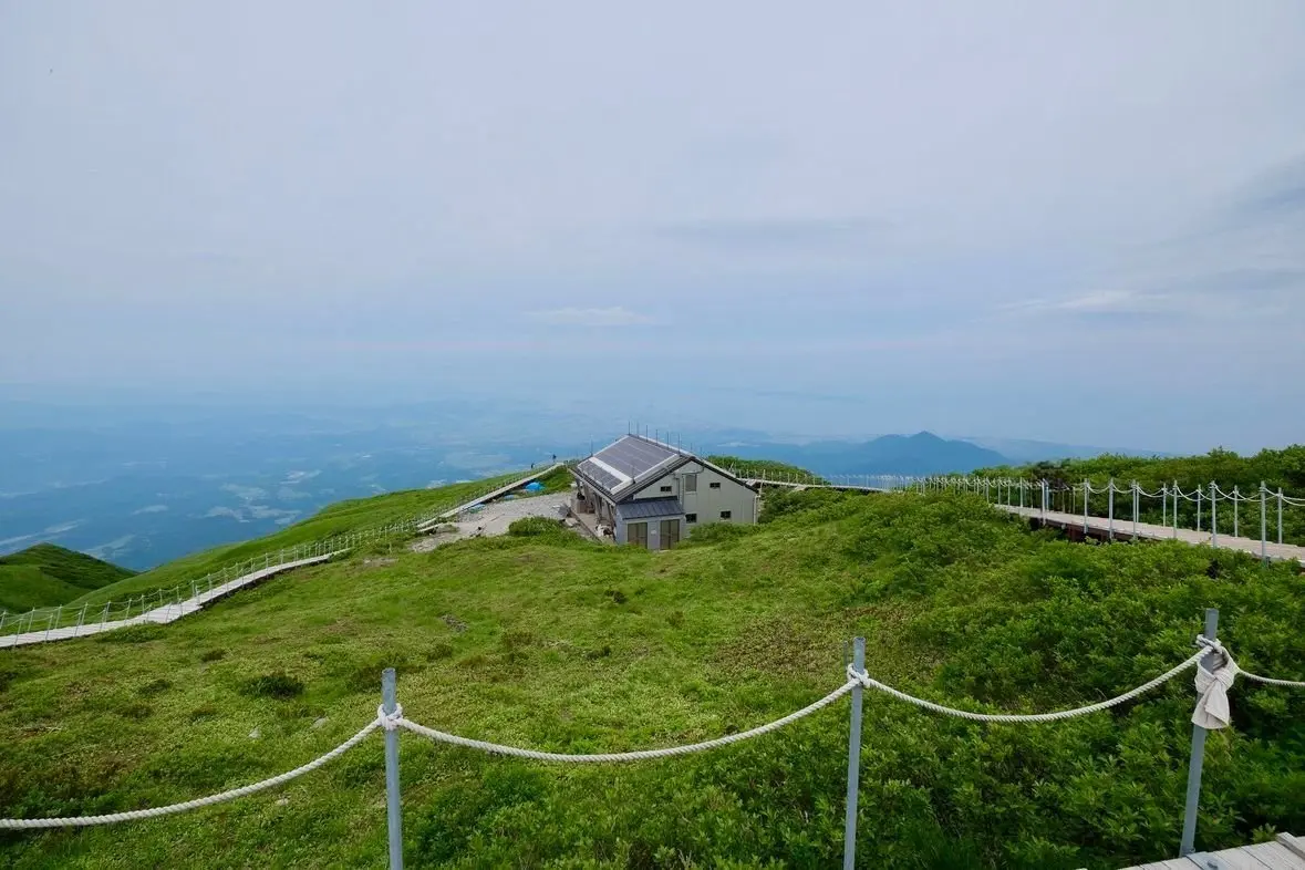 大山の山頂の緑豊かな草原に建つ小屋と手すり付きの歩道、遠くに連なる山々が広がる景色