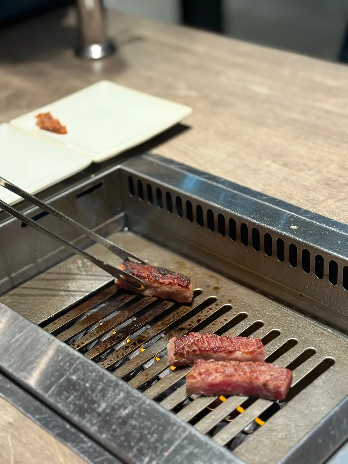 鉄製のグリルで焼かれる牛肉の切り落とし。グリルは格子状で、熱で表面が焼けている。左には白いお皿に盛り付けられた肉片が置かれている。