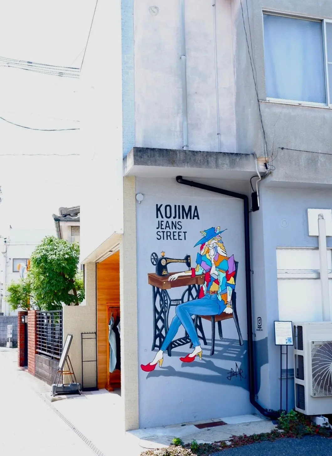 白い壁に描かれた、KOJIMA JEANS STREETと書かれたイラスト。赤い帽子をかぶった女性がミシンに向かい、別の女性が椅子に座って作業している。木製の扉と緑の植物が隣接している。