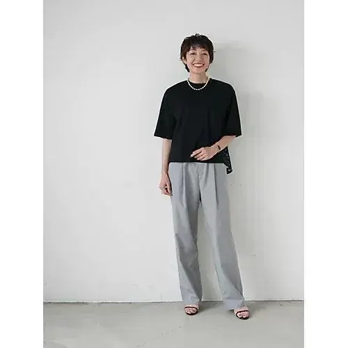 黒いTシャツにグレーのワイドパンツを着用した女性が立っている全身写真。女性は短髪で、白いネックレスとブレスレットを身に着けている。背景は白。