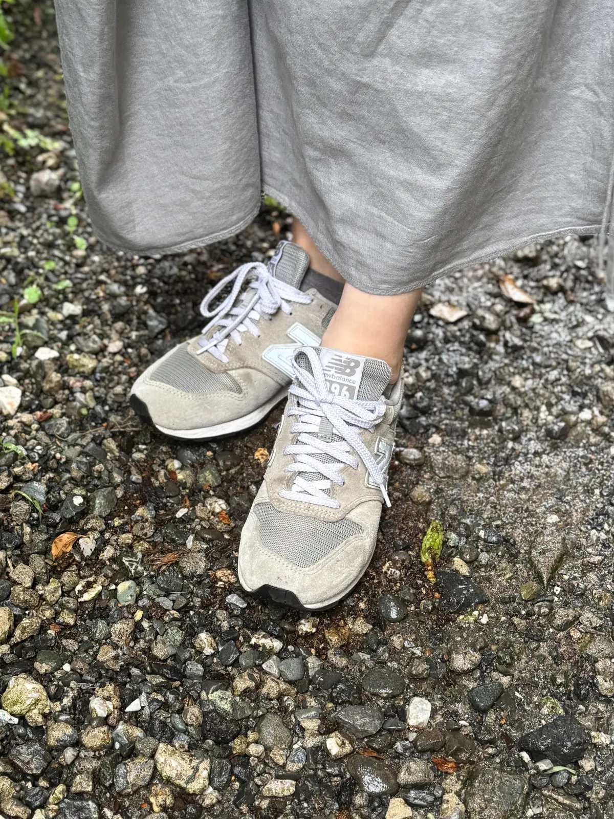 グレーのロングスカートを履いた人物が、ニューバランスのグレーのスニーカーを履いて石畳の上に立っている。靴ひもは白。