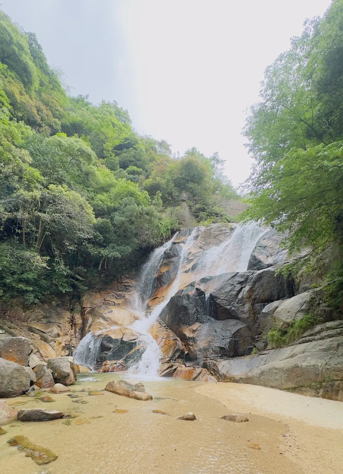 緑豊かな山腹に岩盤が広がり、複数の小滝が流れ落ちる自然の風景。川床には丸い石が散りばめられ、透明な水が岩間を滑る。