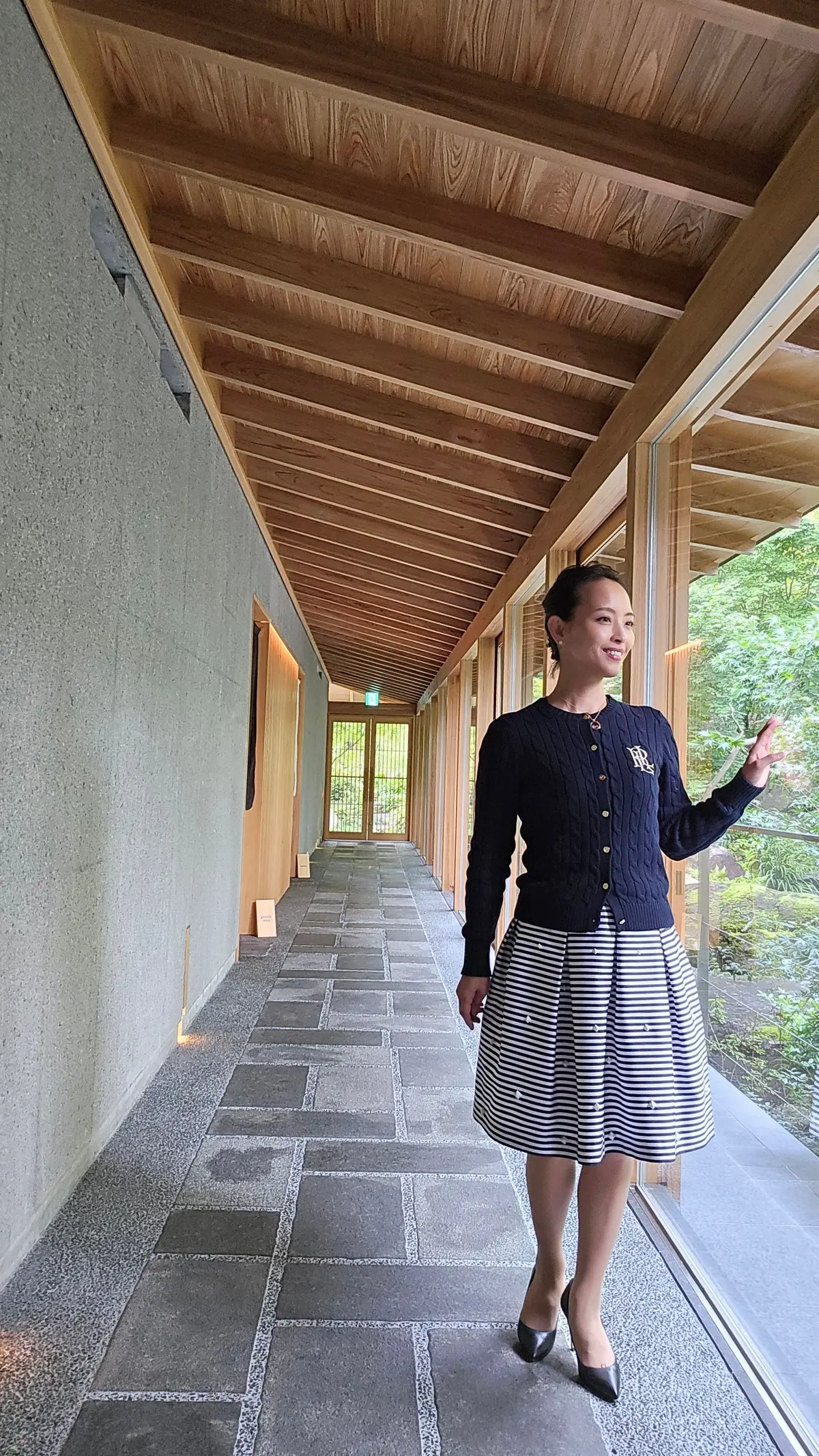 木造の屋根と白い壁が特徴的な回廊に立つ女性。紺のカーディガンに白と青のストライプのスカート、黒いパンプスを着用し、笑顔で手を振っている。背景には緑豊かな庭園が見える。