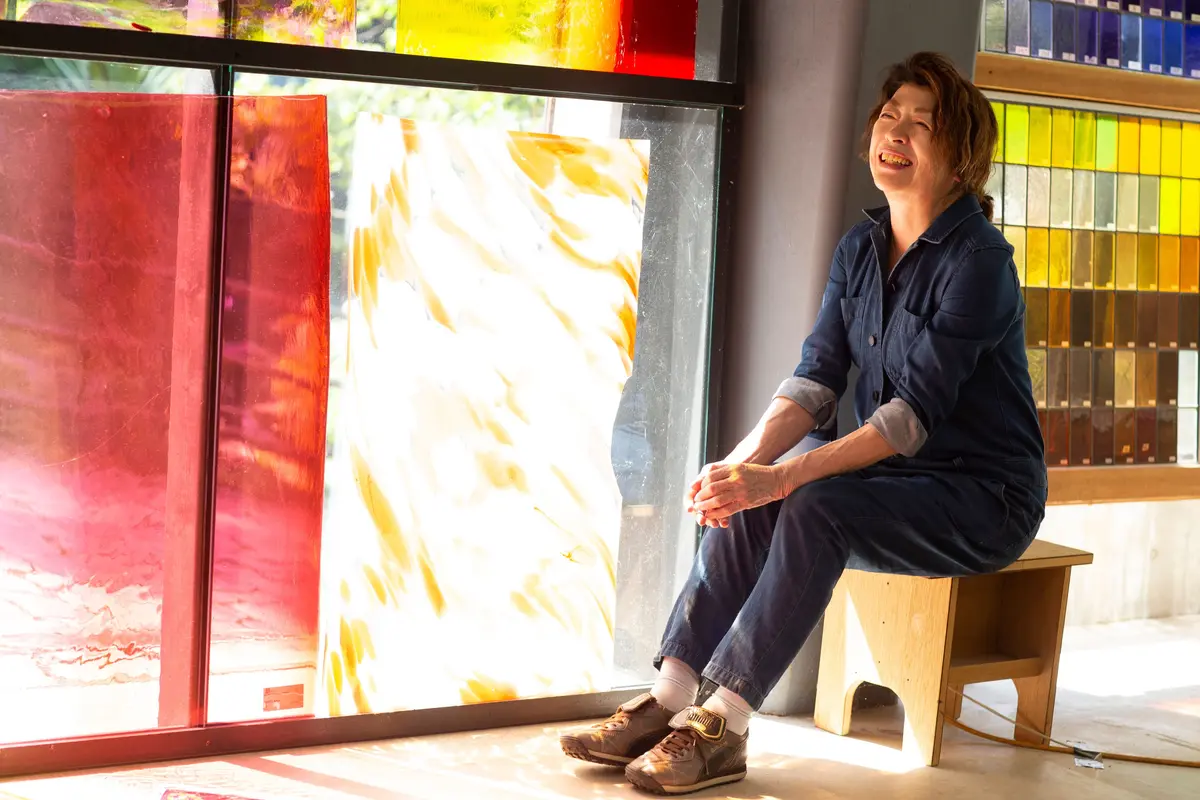 工房でステンドグラス制作をする山本容子さん 笑顔で座り、赤いガラスと淡い黄色のガラスが並ぶ窓の前にいる。右にはカラフルなガラス板が並んでいる