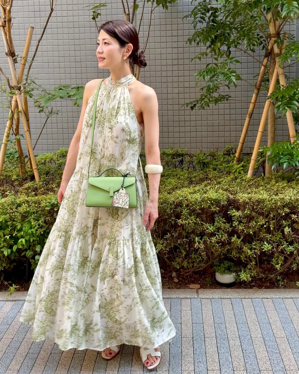 白い花柄のロングドレスを着た女性。緑色のショルダーバッグを肩にかけ、白いサンダルを履いている。背景には緑の植物が生い茂り、グレーの壁がある。