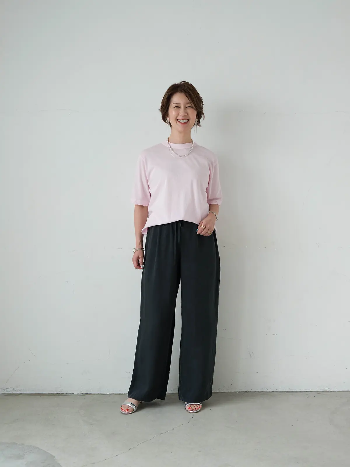 ピンクのTシャツに黒いワイドパンツを着用した女性が、白い壁の前で微笑んでいる全身写真。茶色のショートヘアで、シルバーのネックレスとピアスを身につけている。足元はサンダル。