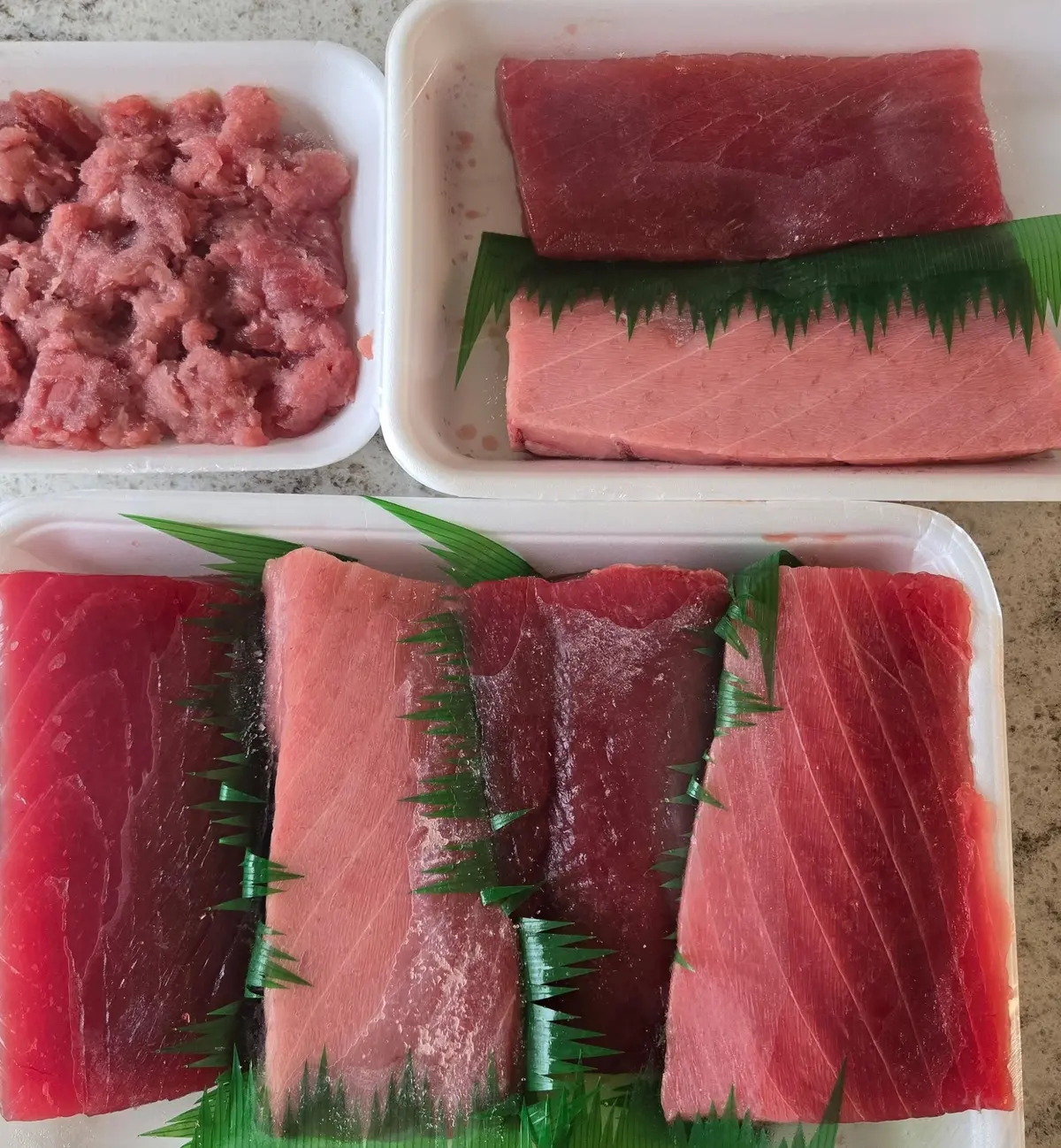 白い木目調の台に、3つの容器に入ったマグロの刺身が置かれている。左上は赤身のミンチ、右上は大きな赤身ブロック、下は薄切りにした赤身の刺身。刺身の間には緑色のネギが挟まっている。容器には「大国屋」のロゴマークが入っている。