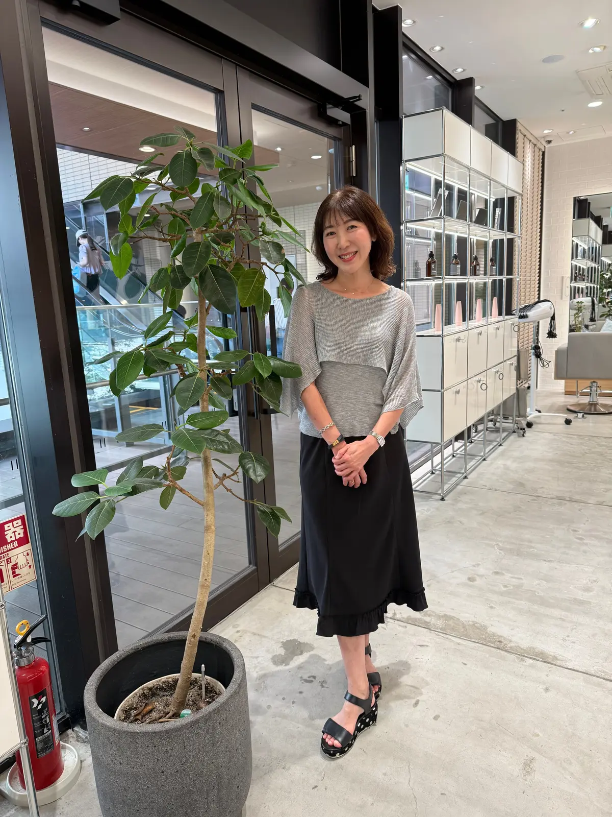 グレーのトップスと黒のスカートを着用した女性が、観葉植物の横に立って微笑んでいる。背景には、モダンなデザインの店内とエスカレーターが見える。