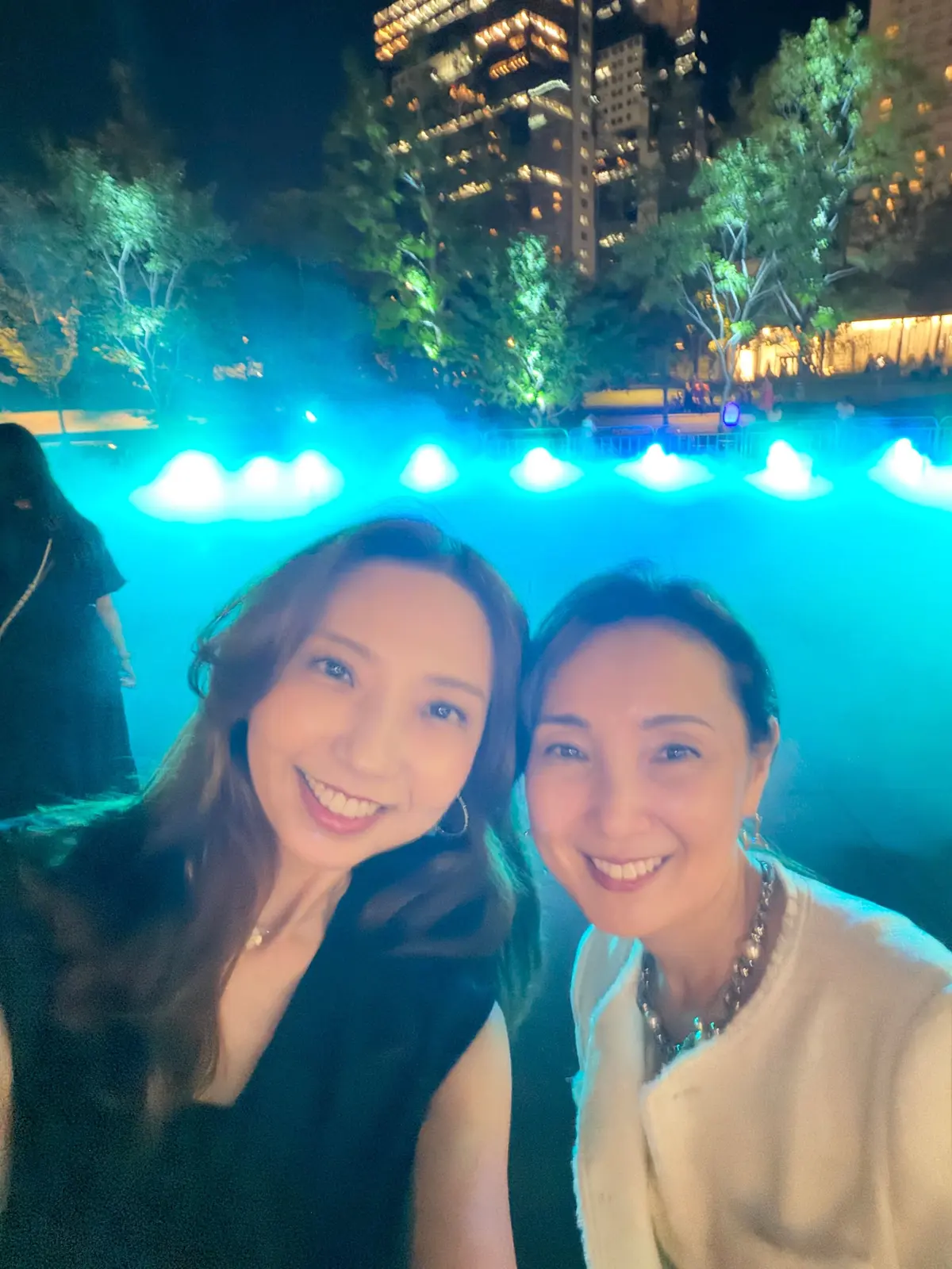夜の公園で、2人の女性が笑顔でセルフィーを撮っている。背景にはライトアップされた水辺と高層ビル群が見える。