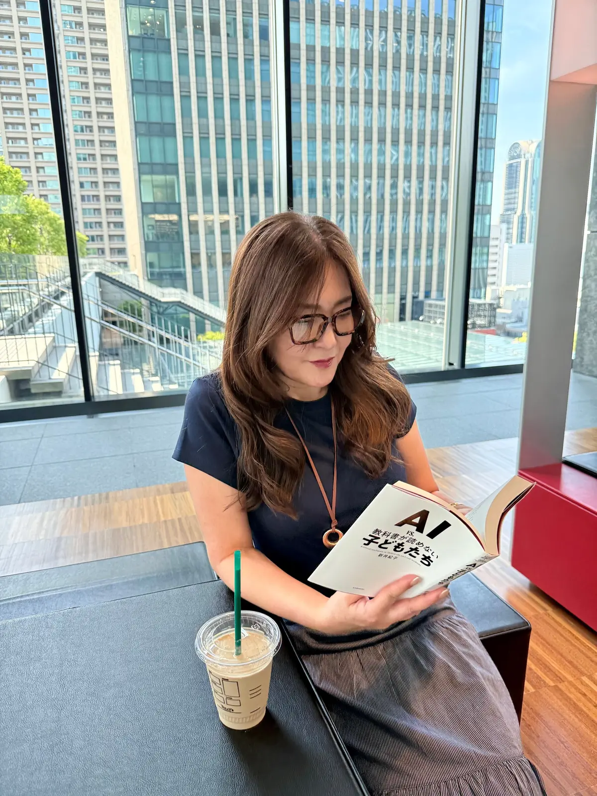 眼鏡をかけた女性が、窓際に座って本を読んでいる。紺色のトップスを着ており、手にはスターバックスのドリンクを持っている。背景には高層ビル群が広がり、窓から光が差し込んでいる。エクラ読書部。エクラ。夏の読書部。おすすめ本。
