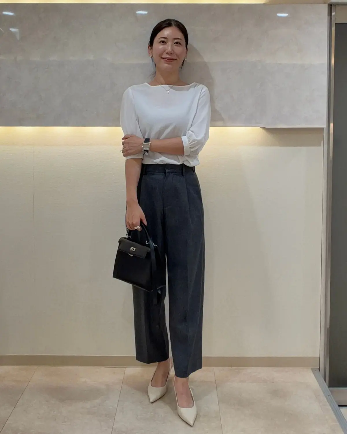 白いカットソーにグレーのワイドパンツを合わせた女性。右手に黒いハンドバッグを持ち、上品な装い。背景はベージュ色の壁。éclat×BRADELIS ブラタンクトップを着用。