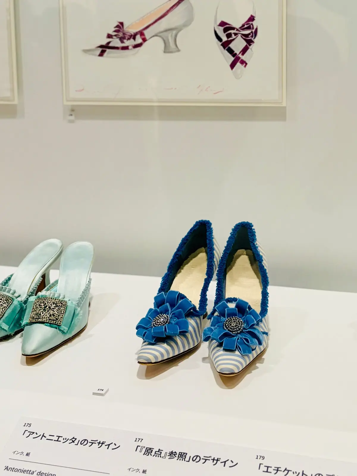 展示ケースに並んだ靴の展示。上部には銀色と紫のリボン装飾が施された靴が2足、下部には青色の装飾花とストライプ柄が特徴的な靴が2足展示されている。それぞれの靴には説明書きが添えられている。