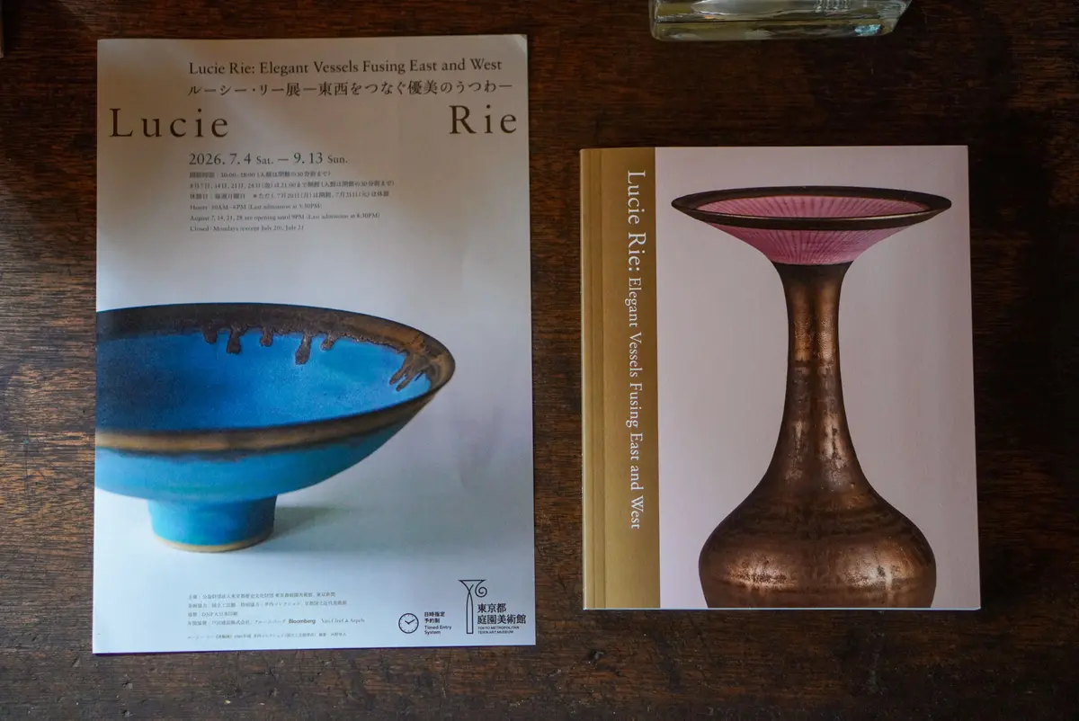 陶芸家ルシー・リーの展覧会ポスター。中央に「Lucie Rie: Elegant Vessels Fusing East and West」のタイトル。背景には、赤茶色と黄色の陶器の作品写真が配置。下部には展覧会の日程や場所などの情報が記載。