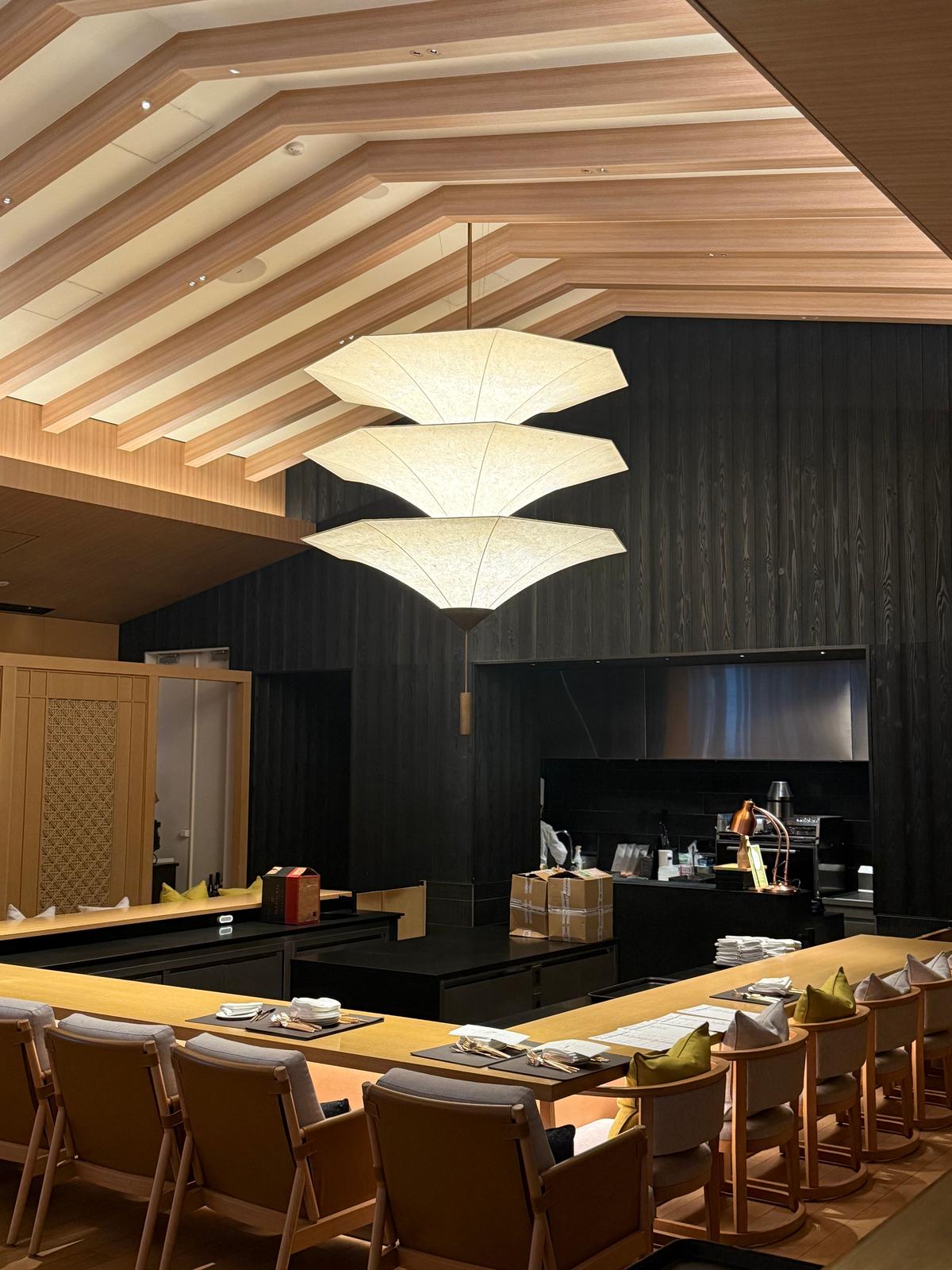 木目調の天井と黒い壁が特徴的な寿司店の内観。中央には傘のようなデザインの大型照明が3連で吊り下げられている。カウンター席には座布団が敷かれ、椅子が並んでいる。背景には厨房があり、調理器具や食材が並んでいる。