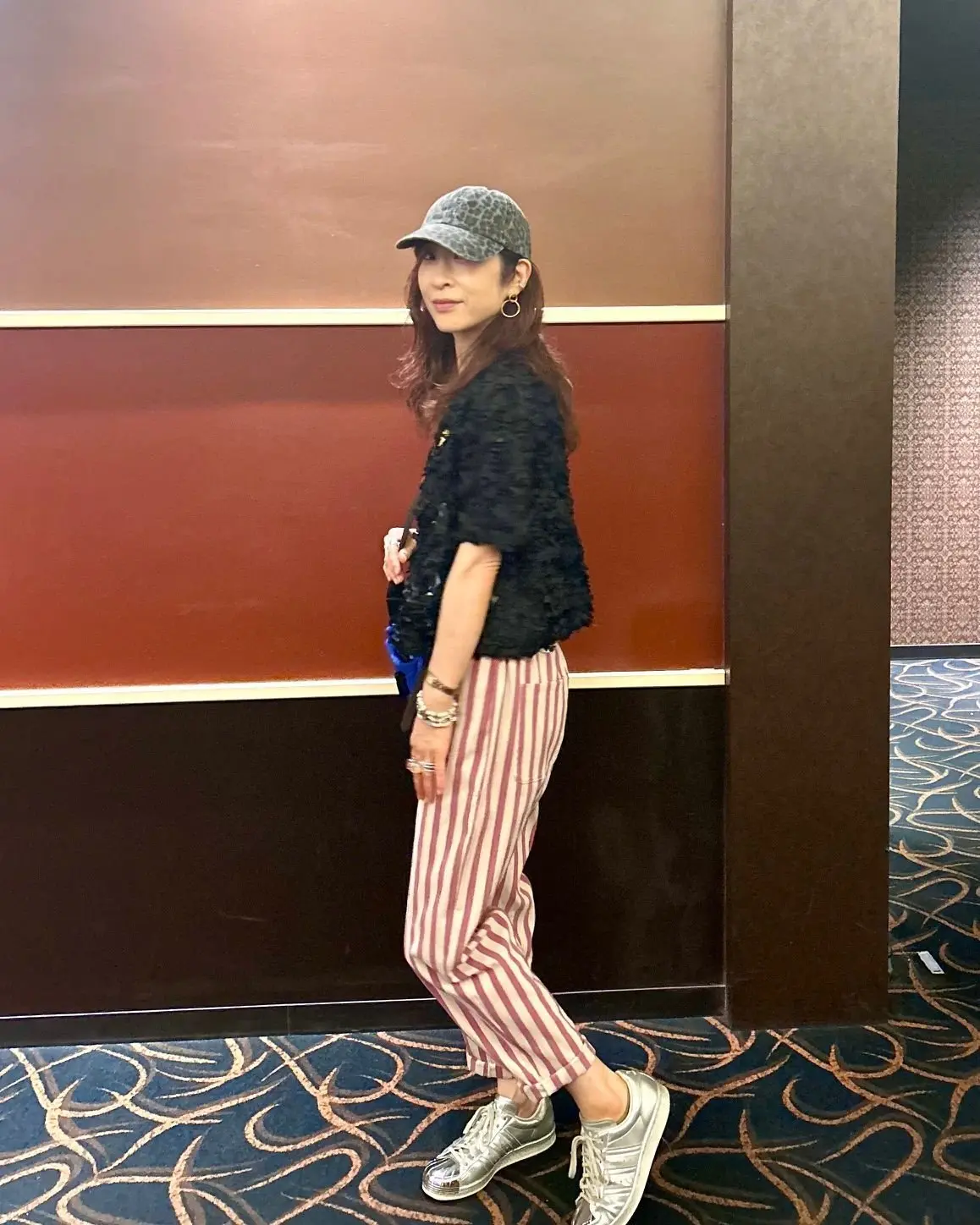女性がキャンバス帽を被り、黒いファージャケット、赤と白のストライプパンツ、銀色のスニーカーを履いて歩く。手には青い小物を持ち、背景に木製の柱と模様のあるカーペットが広がる。