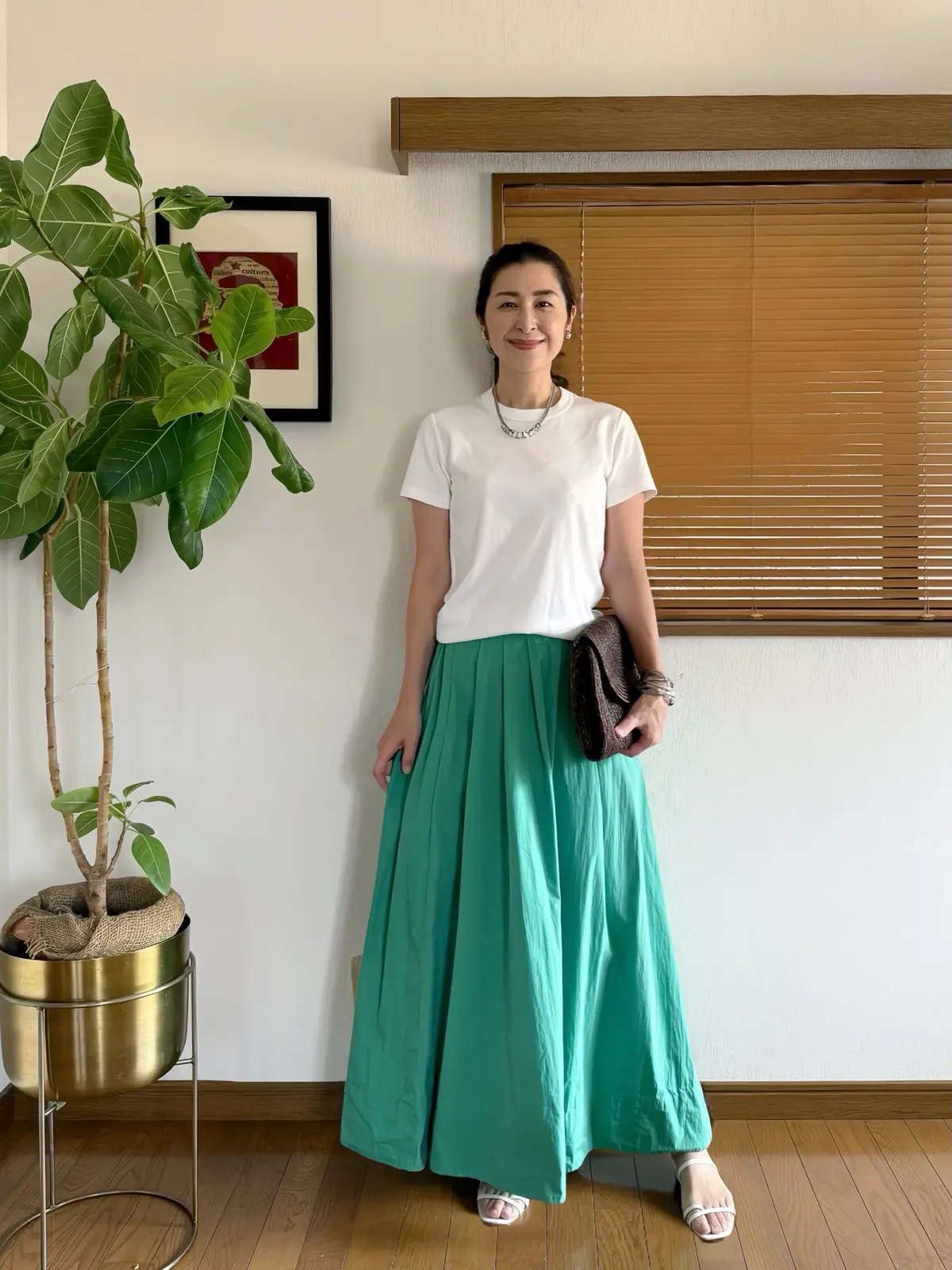 白いTシャツにエメラルドグリーンのロングスカートを合わせた女性。右手に黒いバッグを持ち、背景には観葉植物と木製のブラインドが見える。