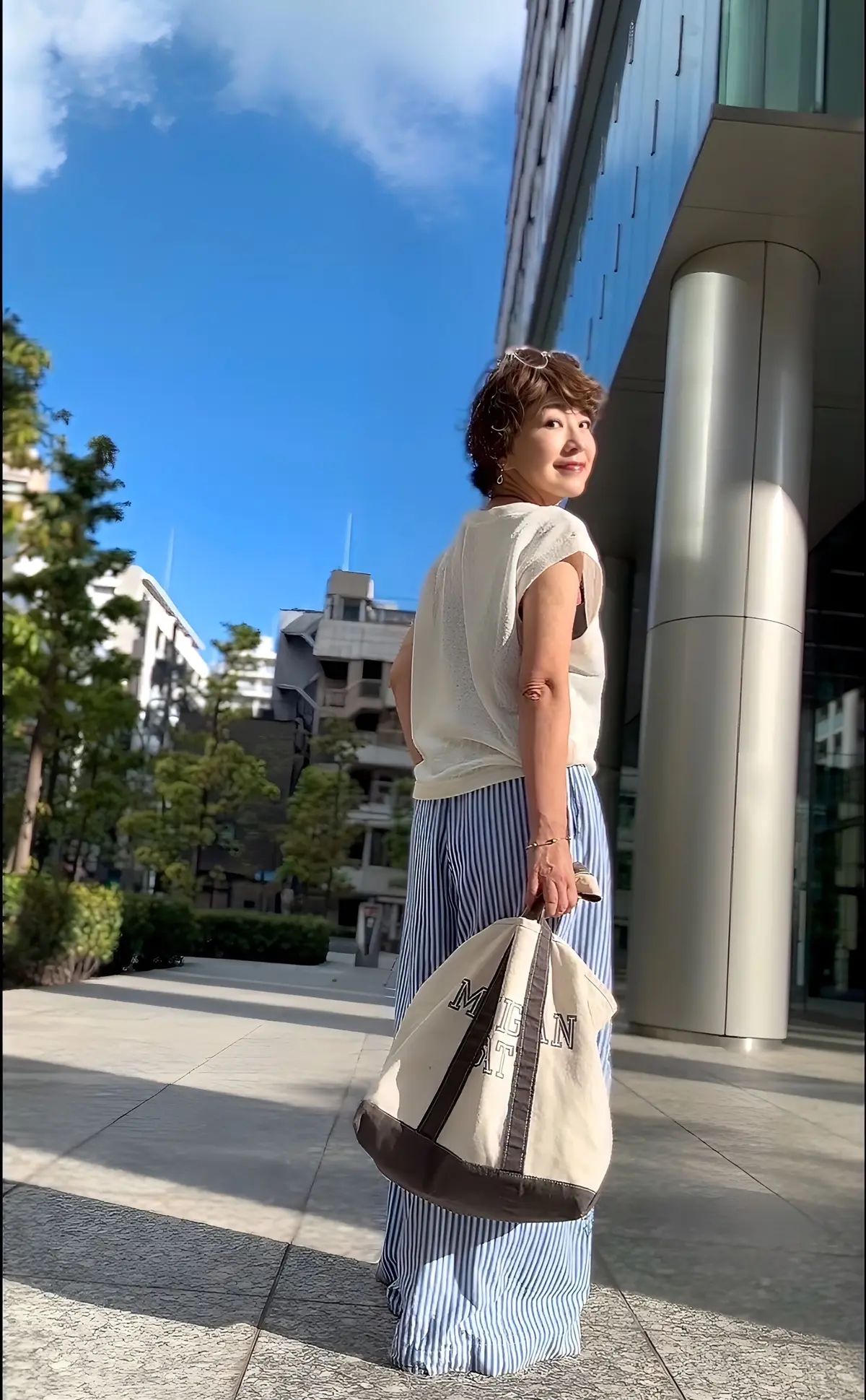 青空の下、白いタンクトップと青白のストライプのパンツを着た女性が、MILANと書かれたトートバッグを持って振り返る。背景には現代的な建物と緑豊かな木々が見える。