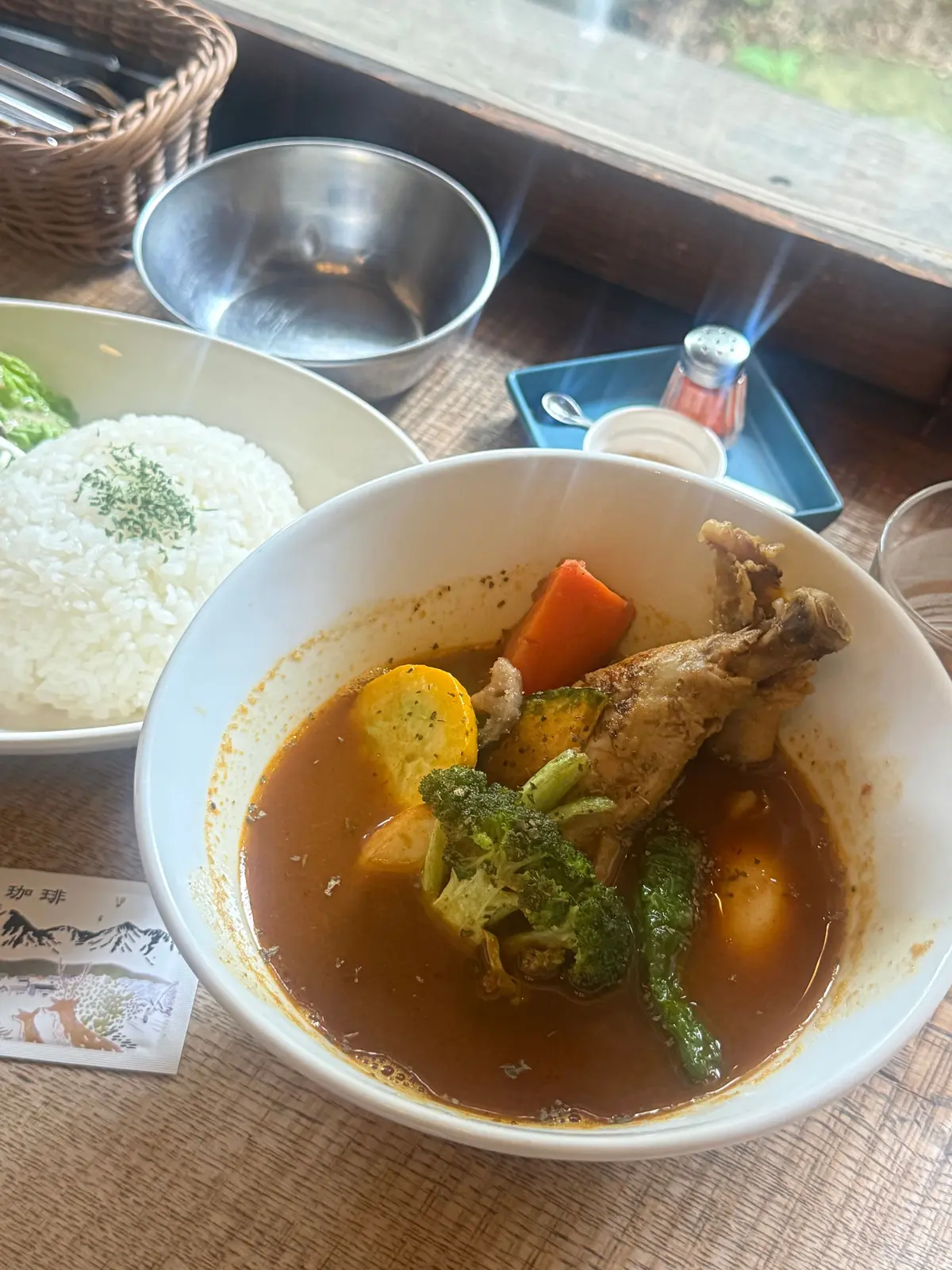 白いボウルに鶏肉、ブロッコリー、にんじん、じゃがいもが入ったカレー風味のスープ、隣には白米と緑の野菜が盛り付けられた料理。