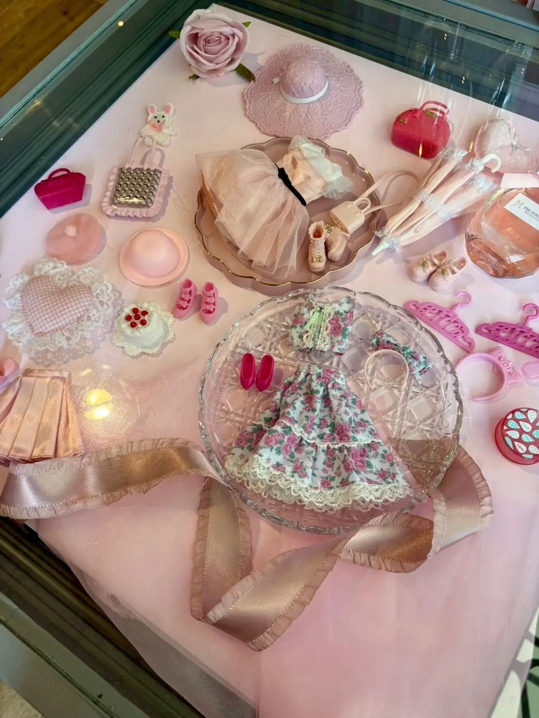 ピンクのテーブルクロスに、ドール用のお洋服や小物、ピンクのハット、靴、アクセサリー、小さなケーキが散らばっている