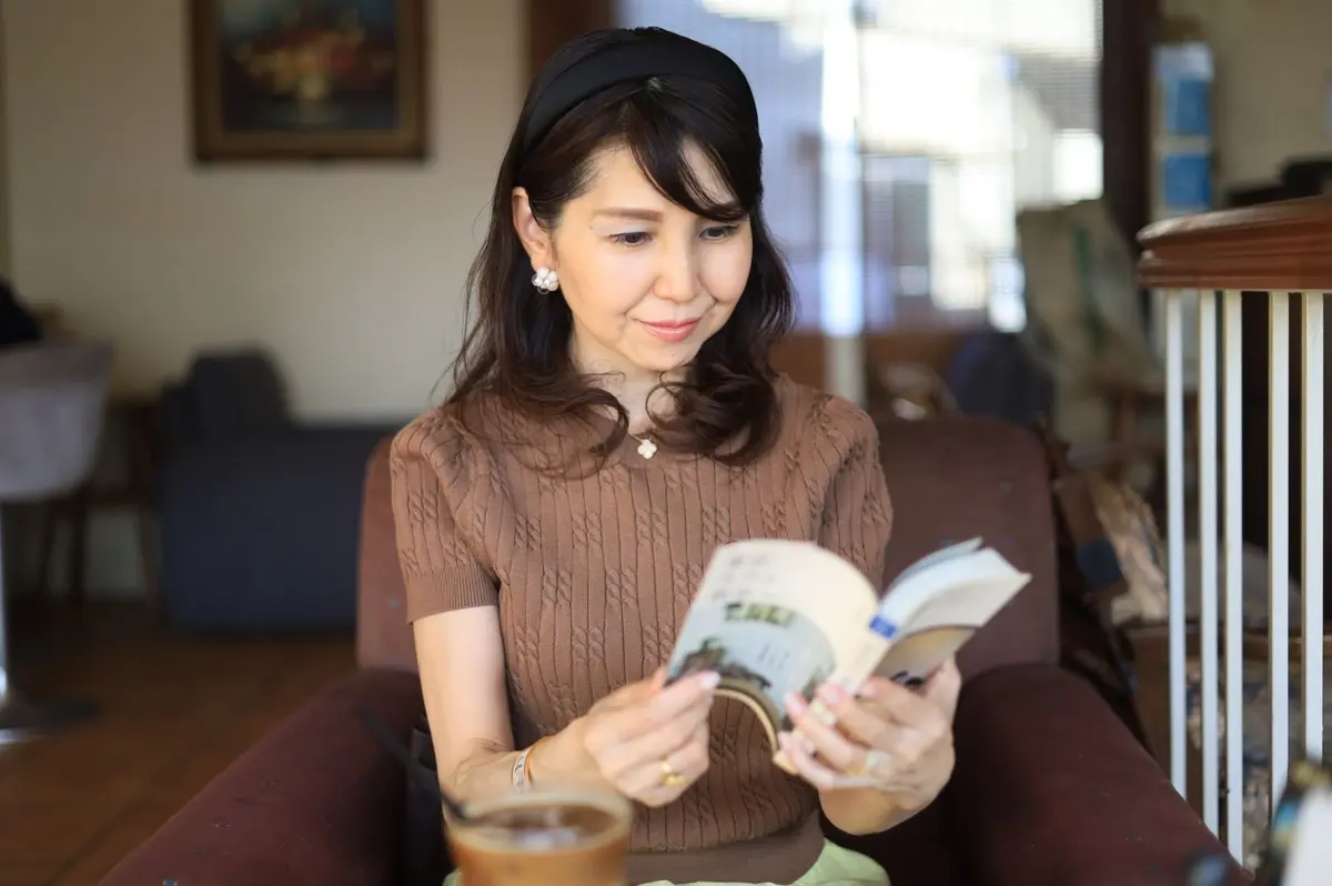 茶色の編み物セーターの女性が、茶色のソファに座り本を読んでいる。手元にはコーヒーカップが置かれている。背景には絵画と窓が確認できる。