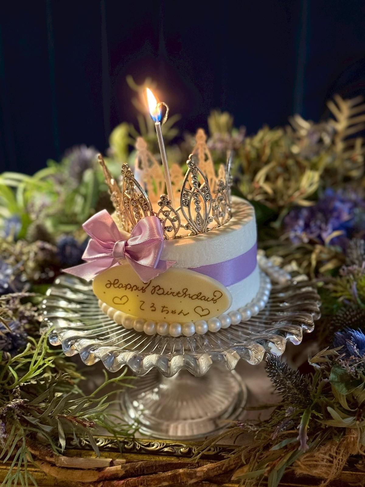 梅田ブルーバードの誕生日サプライズで登場した華やかなティアラケーキ　白と紫の小さなケーキに王冠のトッパーと「ハッピーバースデー」のプレートが乗せられ、キャンドルが点灯している