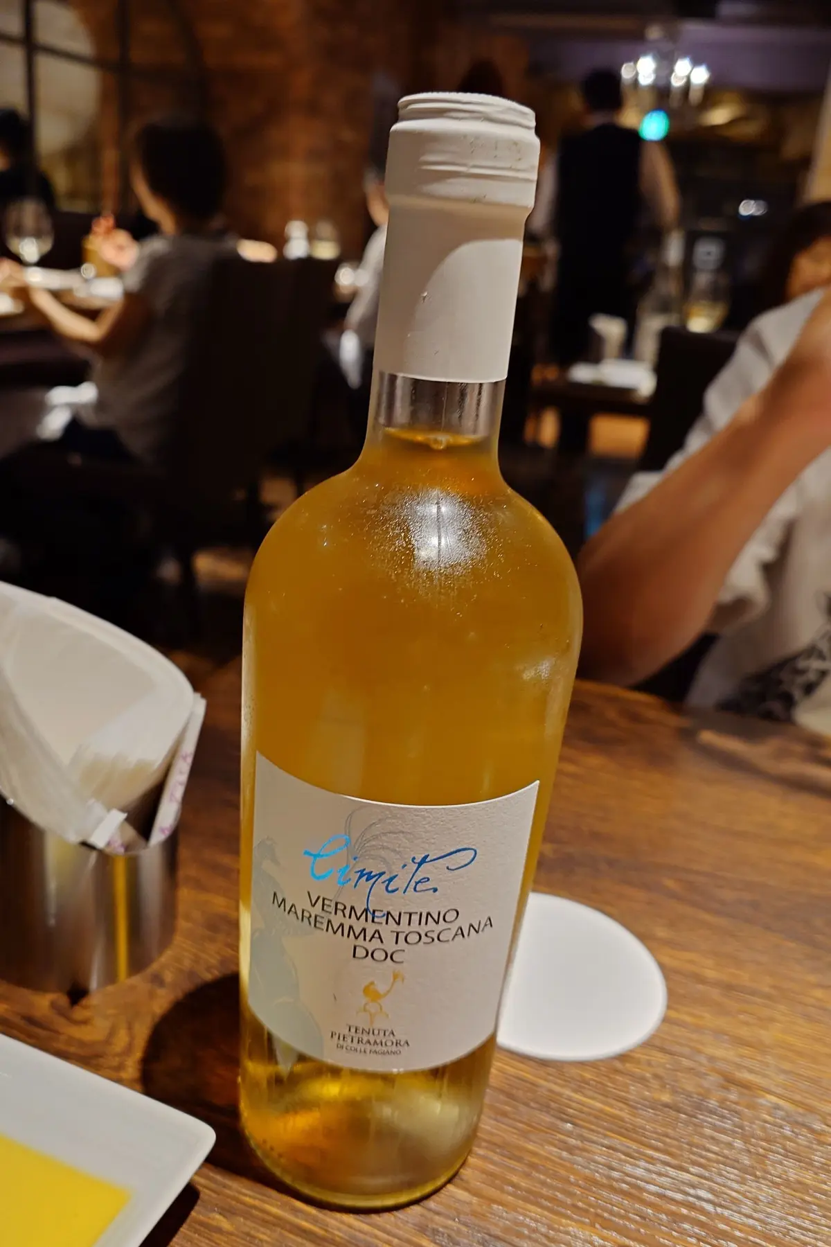 レストランのテーブルに置かれた、淡い黄色いワインボトル。ラベルには青い文字で「LIMITE VERMENTINO MAREMMA TOSCANA」と記載されている。
