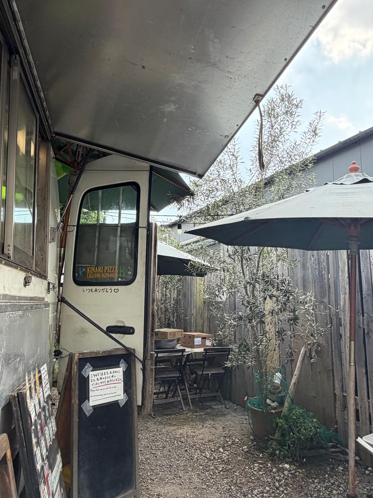 白い箱型のキッチンカー「KINARI PIZZA」が路地に停泊。黒い看板とテーブル、椅子が置かれ、背には緑の植物が生い茂っている。