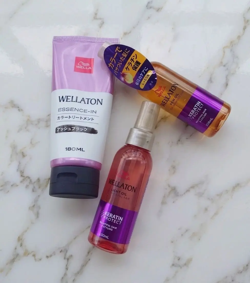 白い大理石の台に置かれた、ウェラ（WELLATON）のヘアケア製品3点。ピンクのチューブ、金色のオイルボトル1本、ピンク色のボトル1本。それぞれ異なる製品名が記載されている。