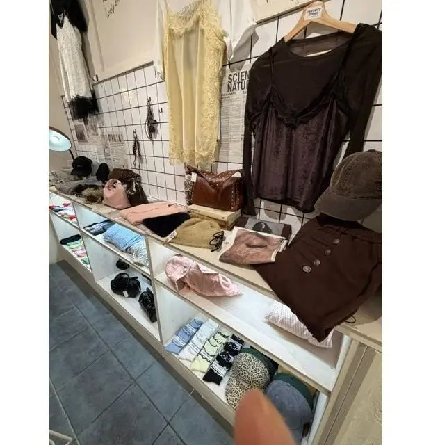 白を基調とした棚に、トップス、ボトムス、アクセサリーなどの衣料品が並んでいる。ブラウンのベルベットトップス、レースのタンクトップ、帽子、バッグ、サングラスなどが置かれている。棚の下段には、柄物のスカーフや靴下などが並んでいる。
