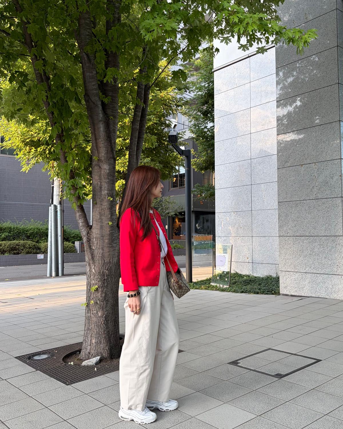赤いジャケットに白いパンツ、白いスニーカーを身につけた女性が、木と建物の前に立っている。女性は横顔でこちらを見ていない。