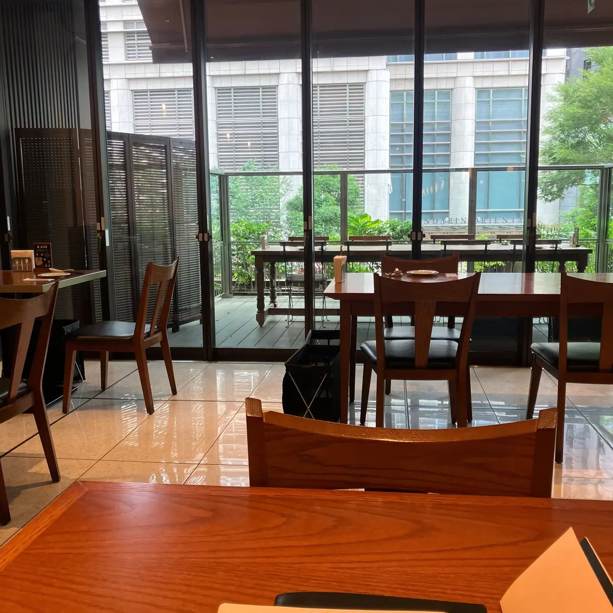 木製のテーブルと椅子が並ぶ明るいカフェの内装。ガラスのドアから外の緑や建物が見え、モダンな雰囲気。