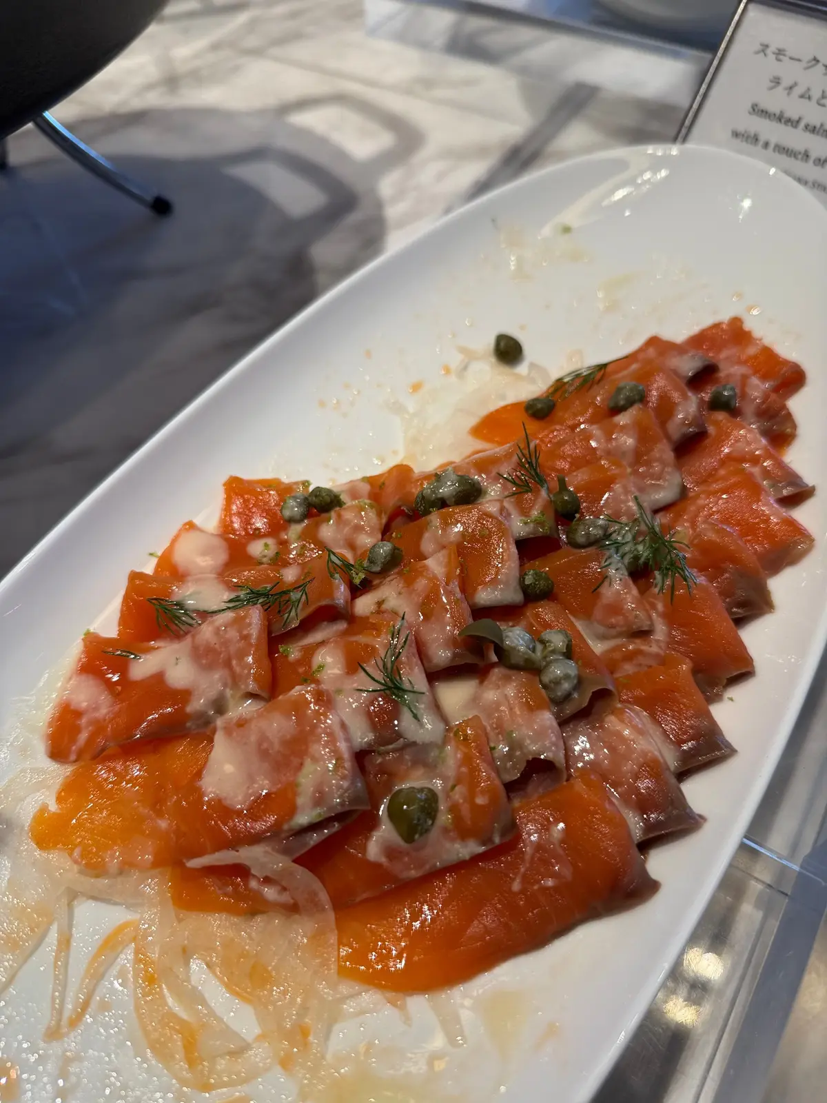 サーモンのスライスにディルとキャップがトッピングされた料理が白い長方形のプレートに盛り付けられています。