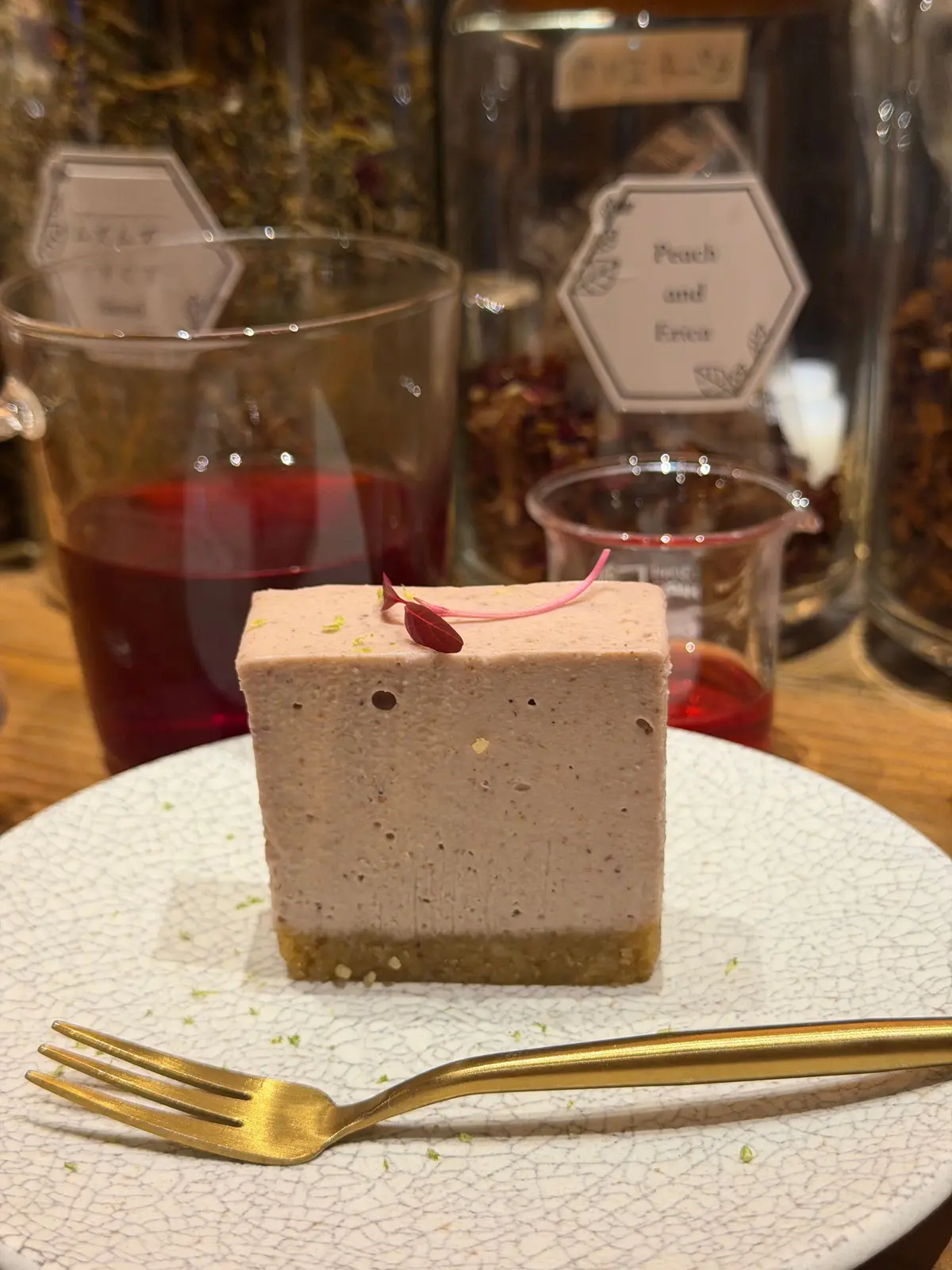 茶色のムースケーキに小さな赤い葉がのったデザートが、クラック模様の白いプレートに置かれています。プレートには金のフォークが添えられ、背景にはハーブの瓶と赤い飲み物が映っています。