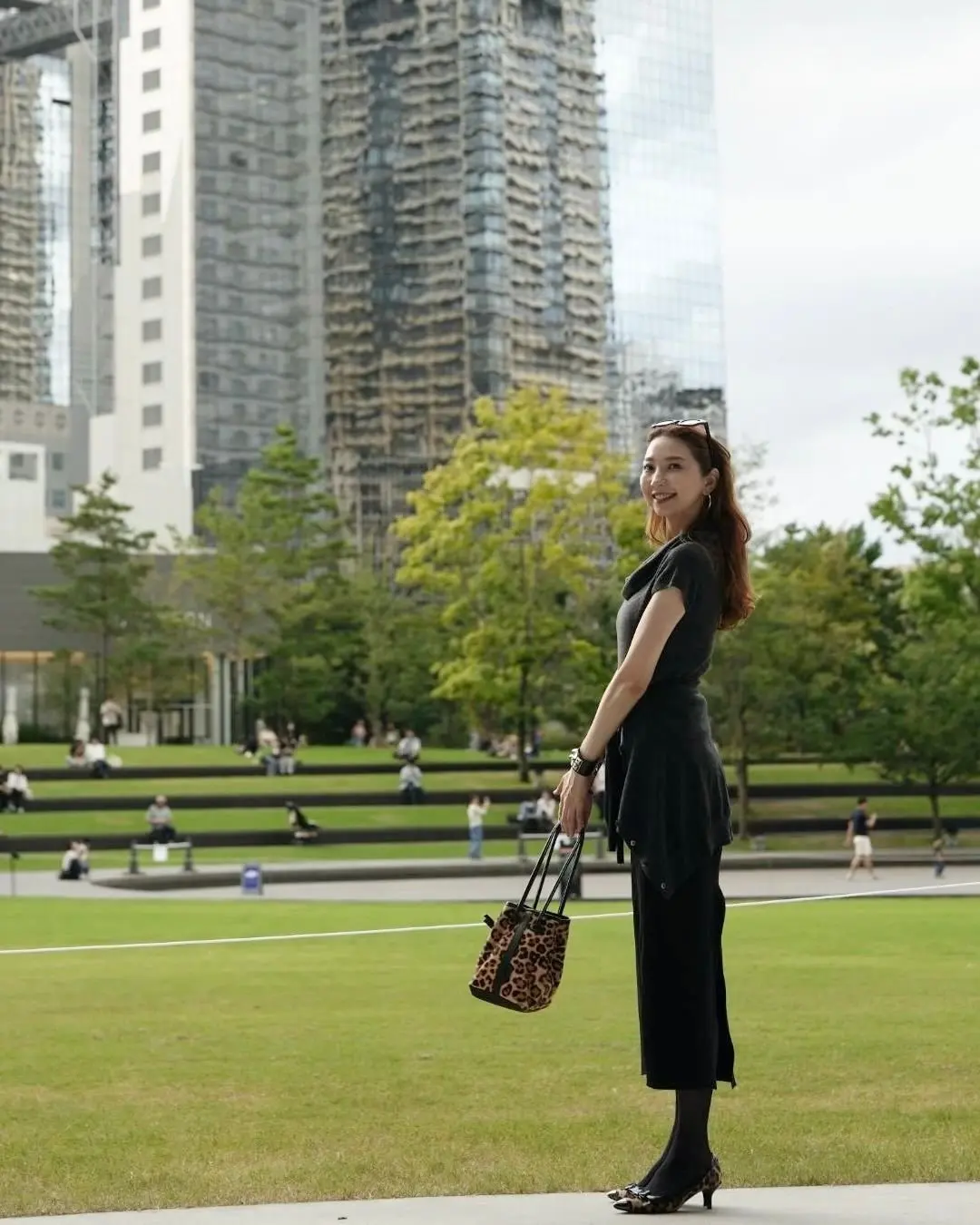 黒いワンピースに身を包んだ女性が、公園の芝生の上で振り返り笑顔を見せている。背景には高層ビル群と、石段状のテラスがある。手にはレオパード柄のバッグを持っている。