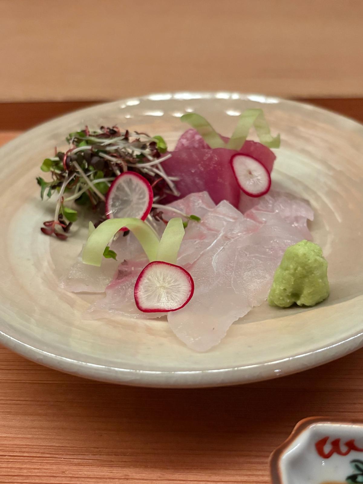 白い器に盛り付けられた刺身。淡いピンク色の魚の薄切りに、赤や緑のスライス野菜、マイクログリーンが添えられている。