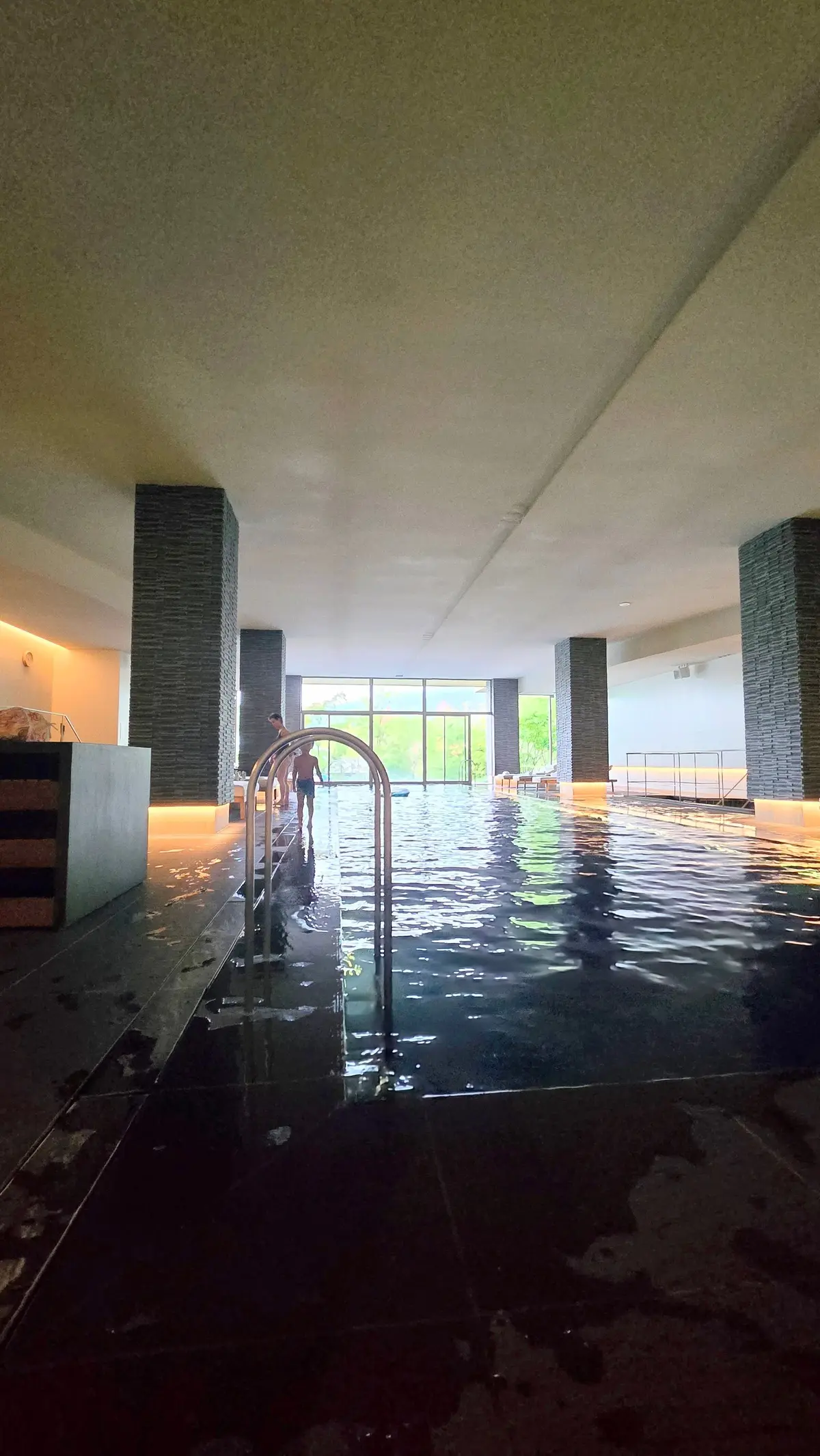 黒色のタイル張りの縁取りと、淡い青色の水面が広がる屋内プール。左右に石柱が並び、奥には窓から緑の景色が見える。手すりが設置されている。