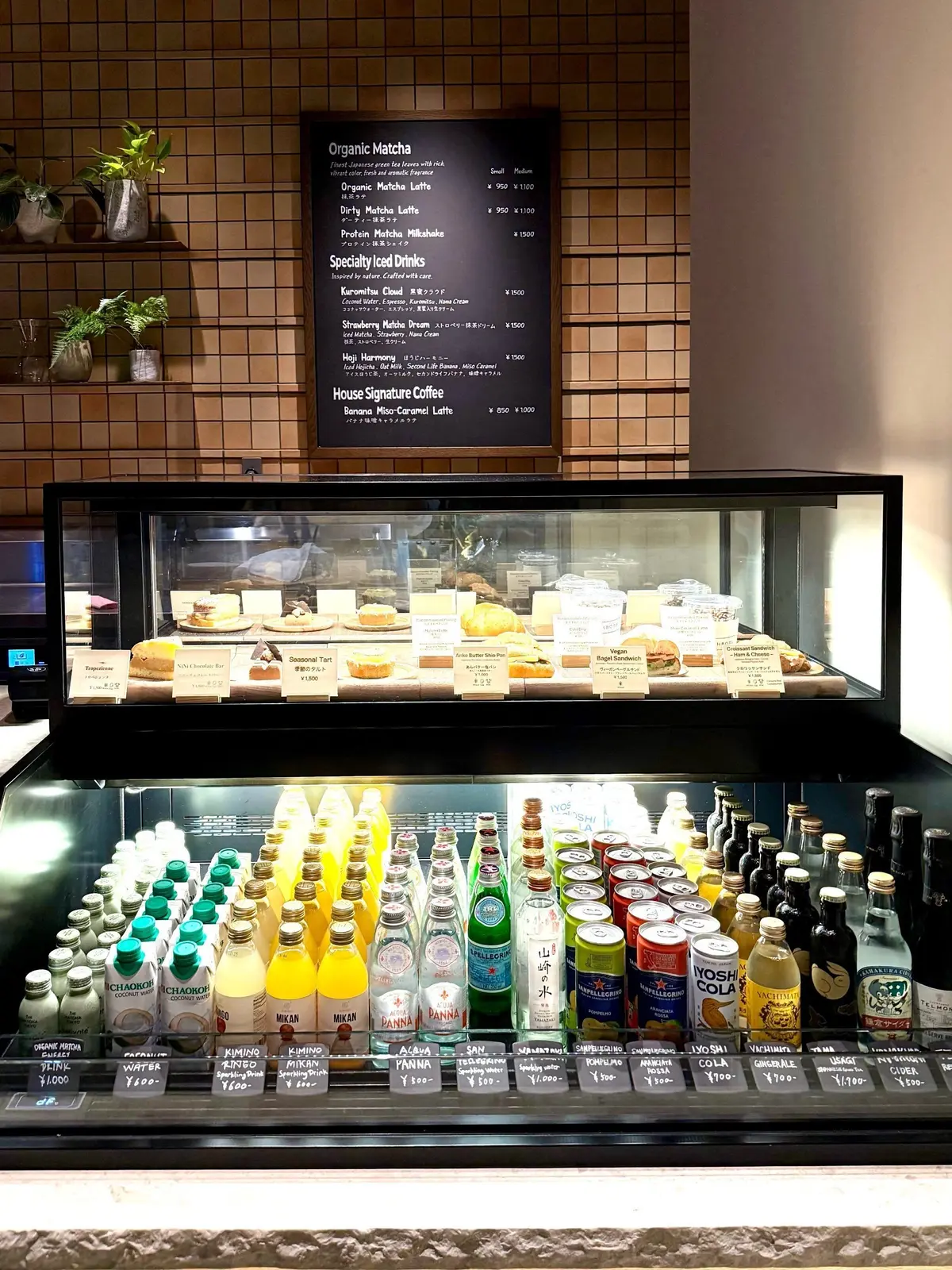 １Hotel Tokyo ３８階　カフェ「Neighbors Café」、カフェのショーケースに各種パンの盛り合わせと多数の缶やボトルの飲料が並んでいる。壁にはマチアやアイスドリンクのメニューが記載されている。