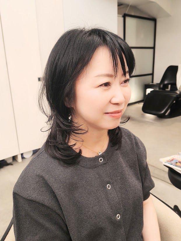 黒髪のボブヘアに前髪を整えた女性が、横を向いて微笑んでいる。グレーのトップスを着用し、ゴールドのネックレスとイヤリングをしている。背景には白い壁と黒い椅子が見える。