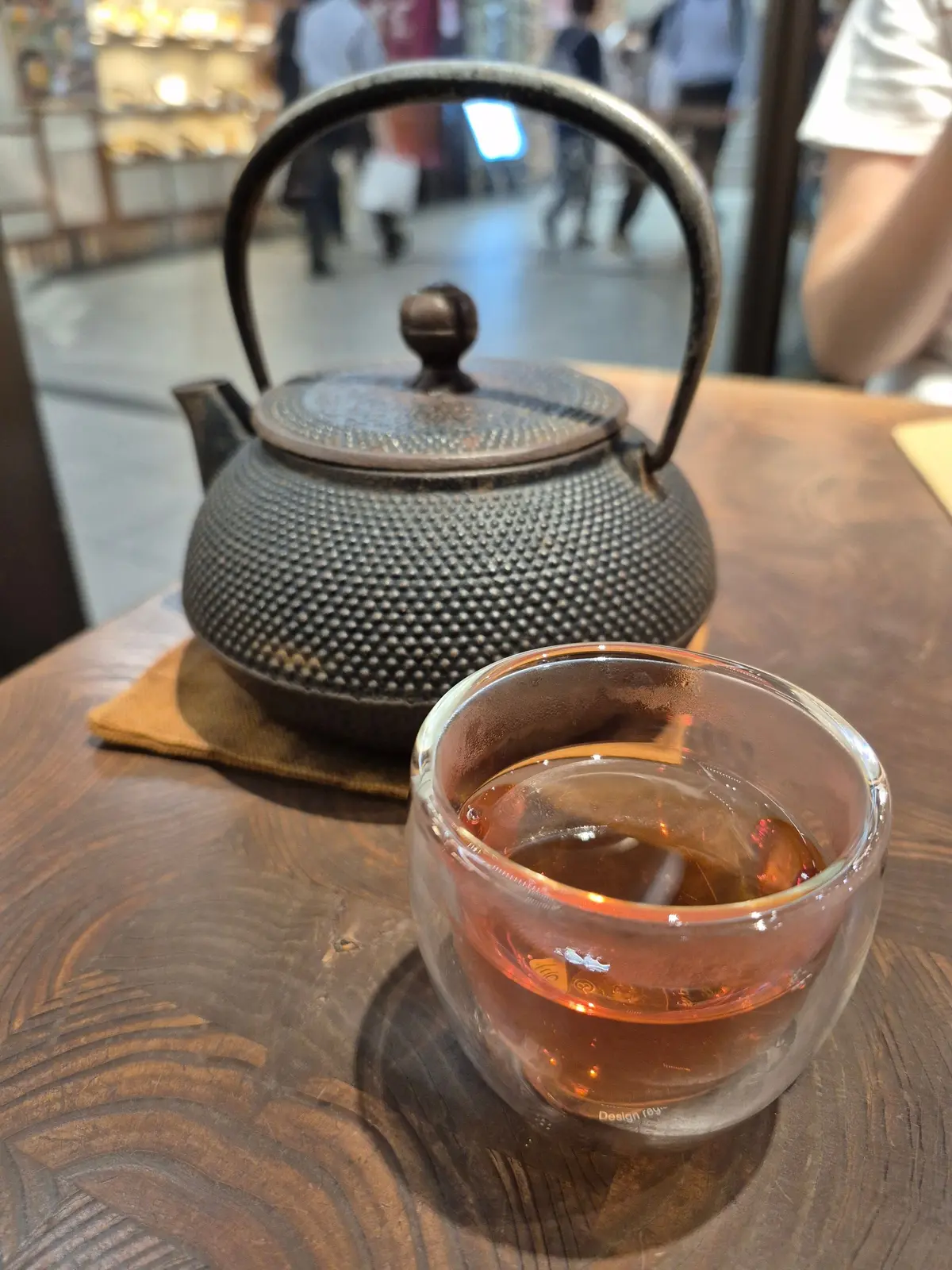 木製のテーブルに置かれた鉄製の茶壺と、ガラス製の湯呑み。湯呑みには琥珀色の飲み物が入っている。背景には人々が歩く店内の様子が見える。
