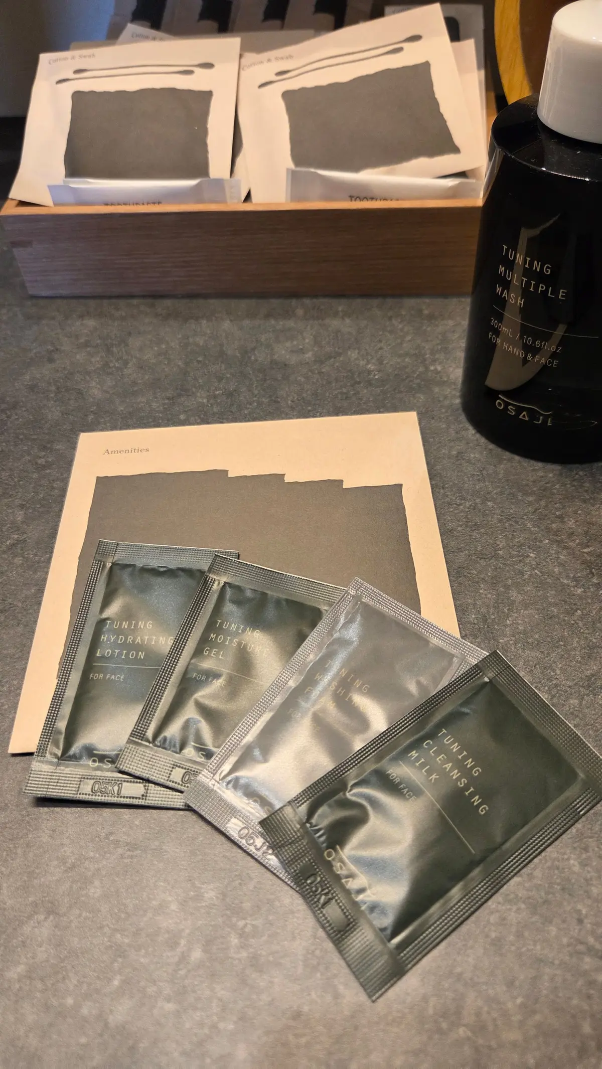 木製のトレイに、OSAJIの洗顔料ボトルと、保湿液、モイスチャーゲル、クレンジングミルクの個包装セットが並んでいる。綿棒やシートマスクも添えられている。