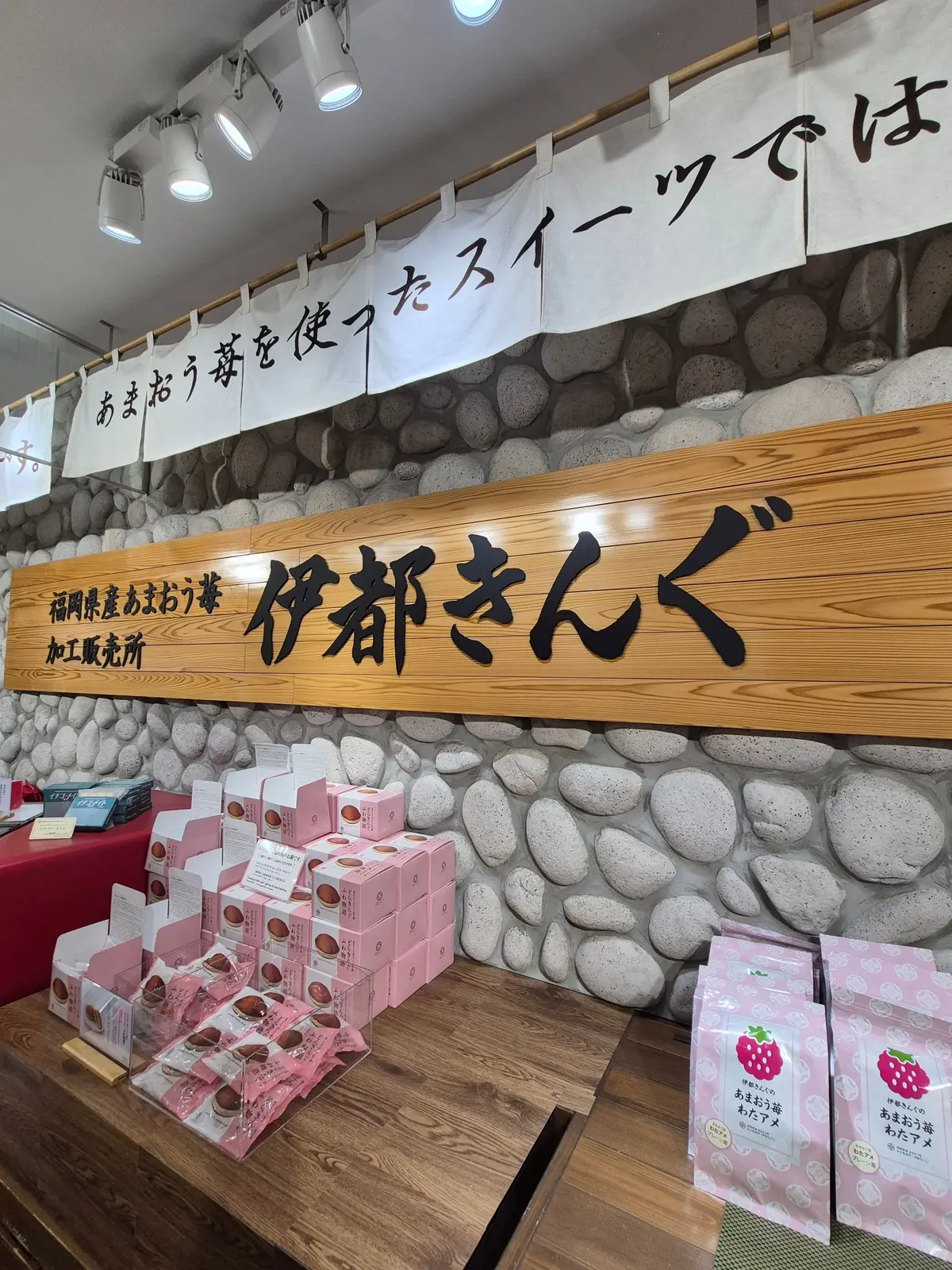 岩を模した装飾が施された壁に「伊都きくら」のロゴがあるお店の様子。木製のテーブルには、ピンク色の箱に入ったお菓子や、イチゴ柄の袋に入ったお菓子が並んでいる。