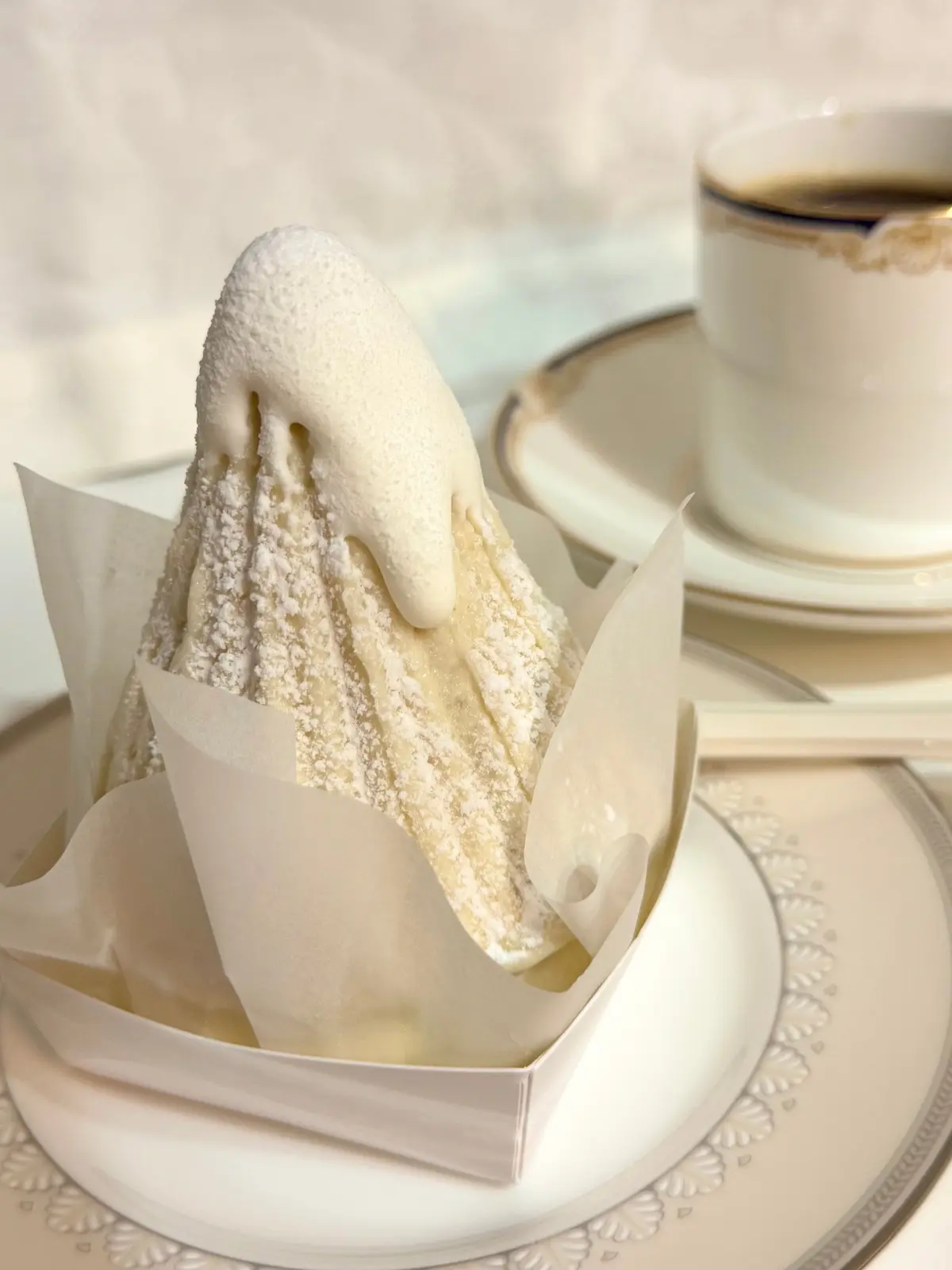 粉砂糖をまぶしたケーキの一切れを金のフォークで持ち上げる、装飾された白いプレート上のケーキとコーヒーカップが並ぶテーブル