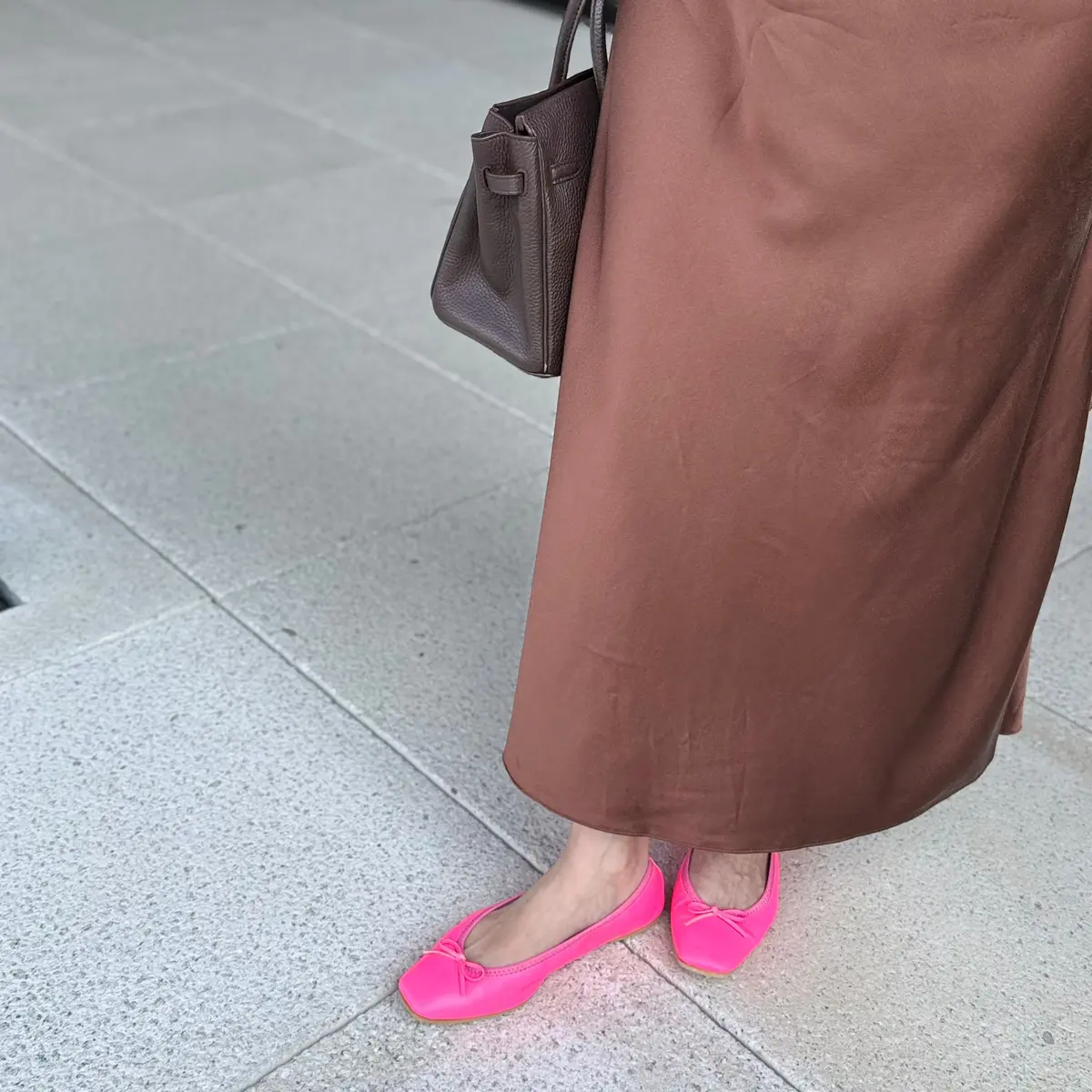 茶色のロングスカートとピンクのフラットシューズを履いた女性。グレーのハンドバッグを肩に下げ、コンクリートの地面に立っている。