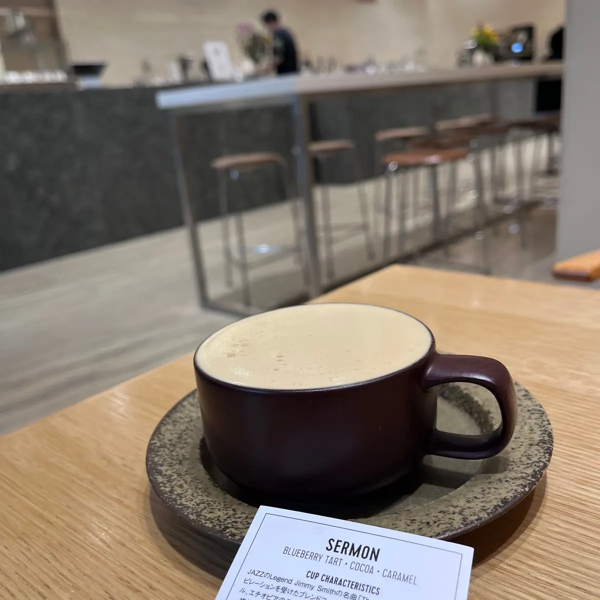 木製のトレーに置かれた、茶色のカップに入ったコーヒー。カップの表面には白いミルクフォームが均一に広がっている。背景には、カフェのテーブルと椅子が見える。