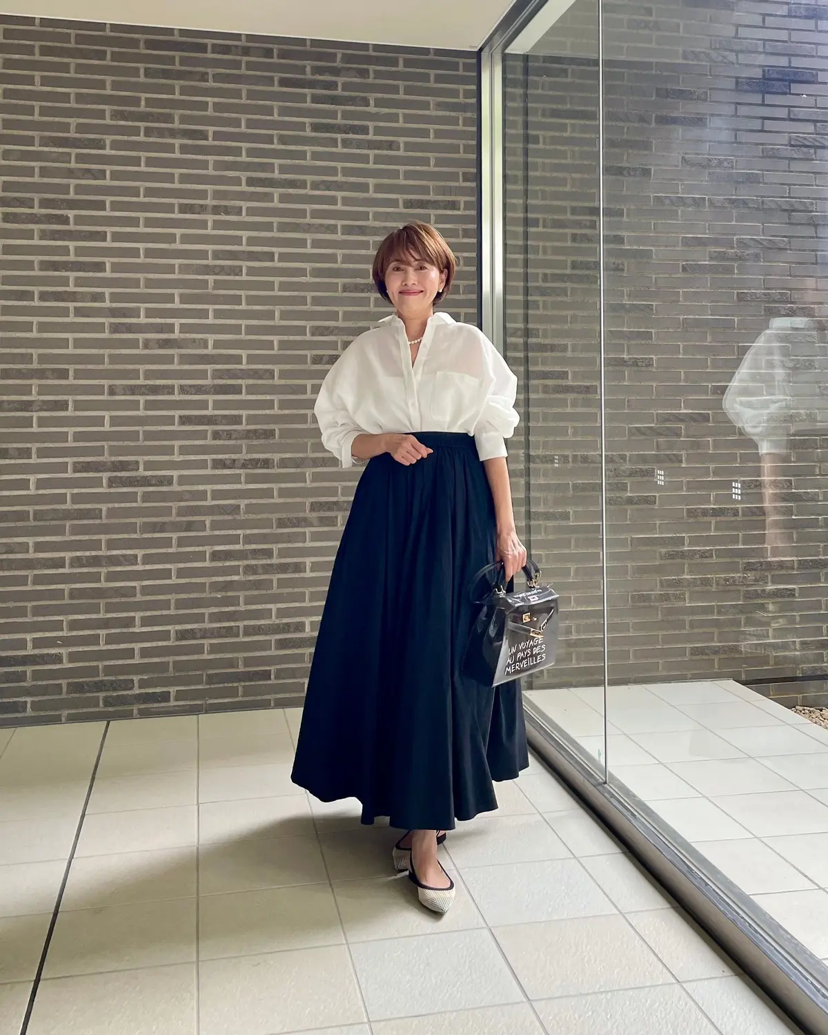 白いブラウスと紺色のロングスカートを着た女性が、ガラスの壁の前に立っています。女性は黒いハンドバッグを右手に持ち、笑顔でカメラを見つめています。背景にはレンガ調の壁が広がっています。