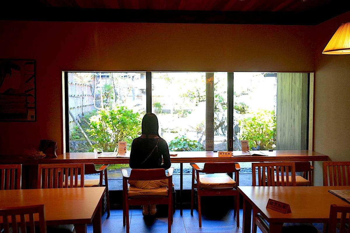 木造の店内、窓際に座る女性。黒い服を着て、こちらを向いていない。窓の外には庭が見え、木々や竹垣が茂る。テーブルと椅子が配置され、温かい照明が灯っている。