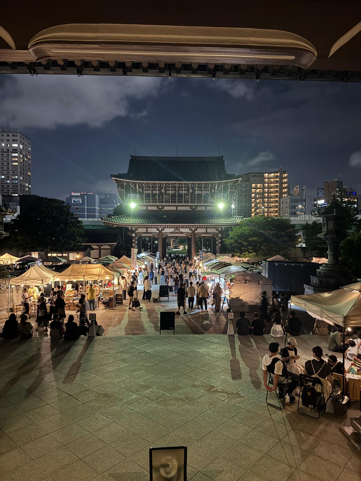 夜の市場の賑わい。伝統的な屋根の建物を中心に、多くの人々が露店を囲み、食事を楽しんでいる様子。背景には高層ビル群が立ち並ぶ。