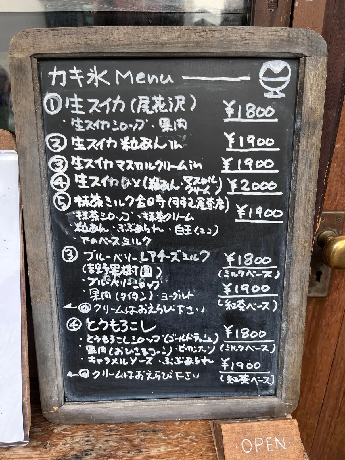 カキ氷のメニューが書かれた黒板。手書きで各メニュー名、価格、説明が記載され、上部にカキ氷のイラストがある。メニューは、生カキ氷、抹茶ミルク、フルーツを使ったものなど、様々な種類が並んでいる。下部には「OPEN」の文字が見える。
