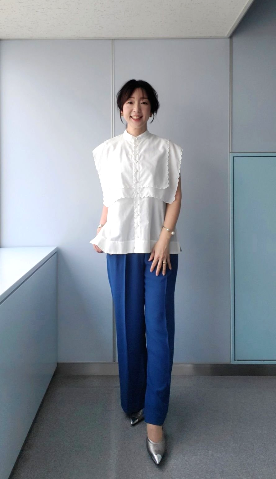 白のフリル付きブラウスと青いワイドパンツを着た女性が、シンプルな室内空間で微笑んでいる。