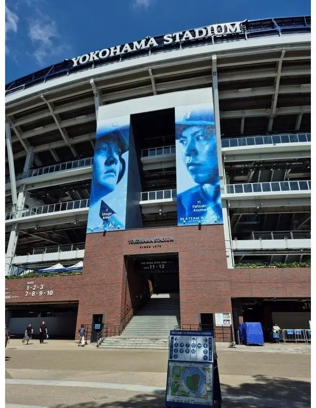 横浜スタジアムの正門付近。赤レンガの建物に、青を基調とした選手2人の巨大なポスターが掲示されている。背景にはスタジアムの座席席が広がり、青空の下、一部雲が見える。